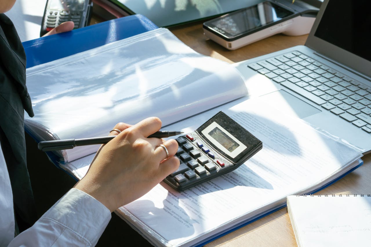 A person working on financial calculations using a calculator and laptop at an office desk.